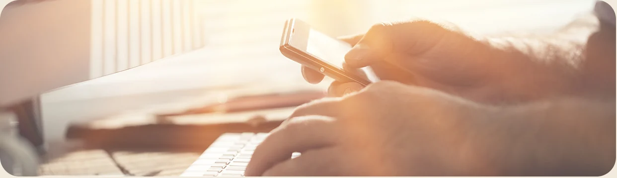 Someone typing at a keyboard while holding a mobile phone
