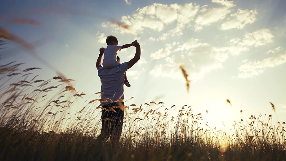 A parent carrying their child on their shoulders in a sunlit field